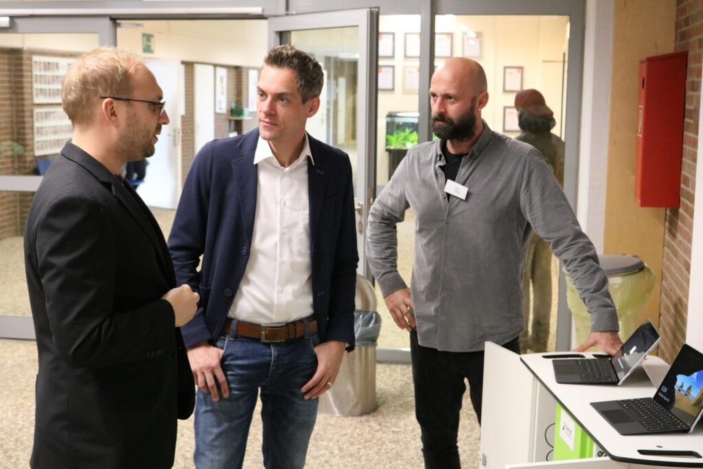 Drei Männer stehen an einem Tisch mit Laptops in einer Schulumgebung.
