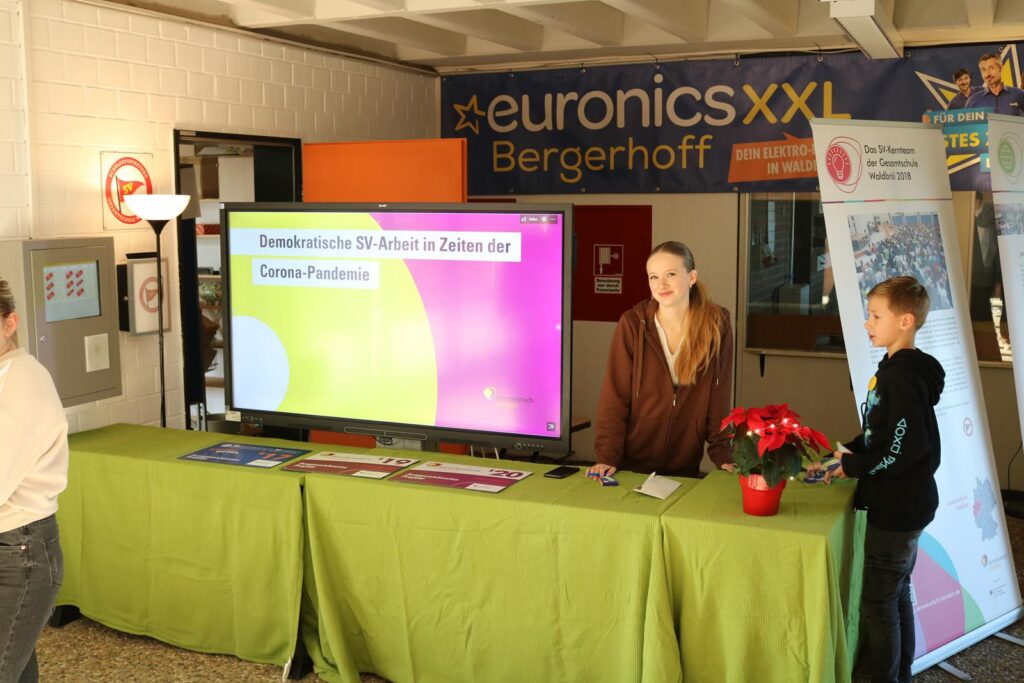 Schülerin am Informationsstand mit Bildschirm und Pflanzen.