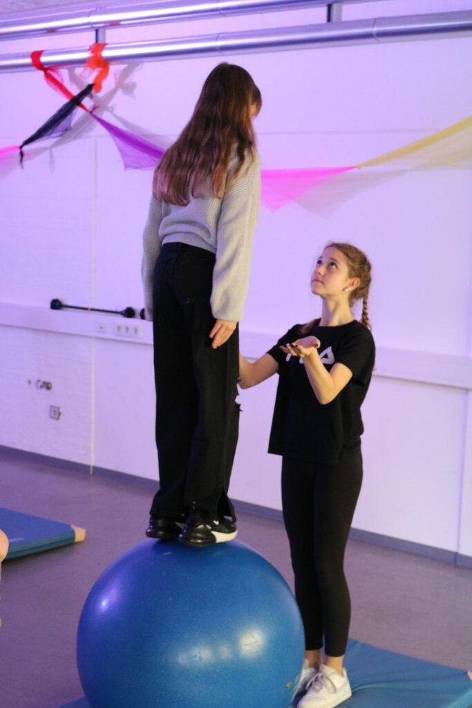 Mädchen steht auf einem Gymnastikball, ein anderes Mädchen gibt Unterstützung.