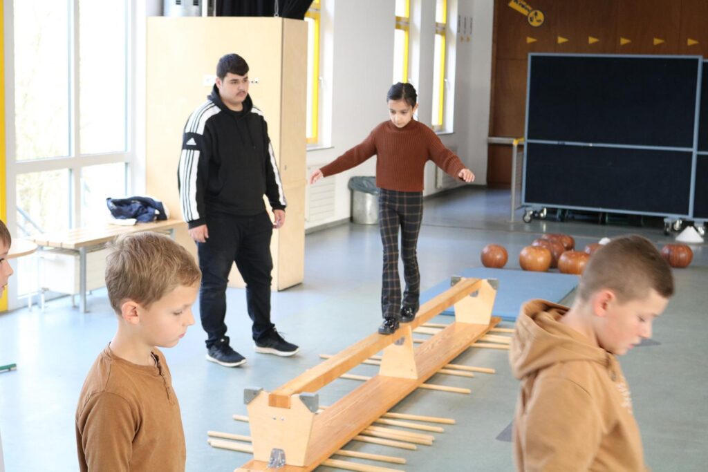 Kinder balancieren auf einem Holzsteg in einer Sporthalle.