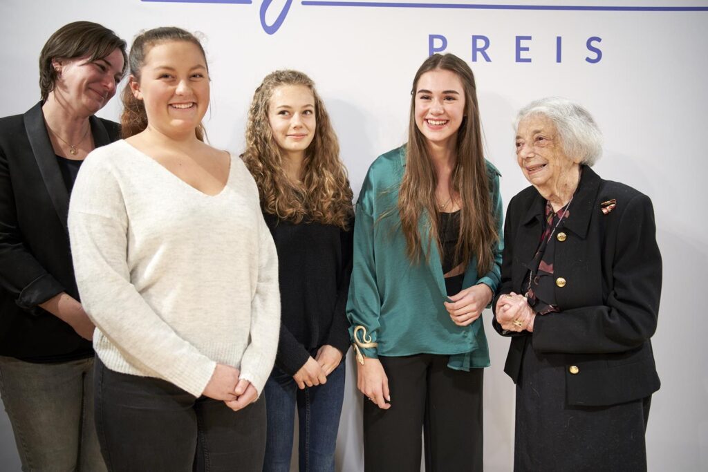 Fünf Frauen stehen lächelnd vor einer Wand mit Text.