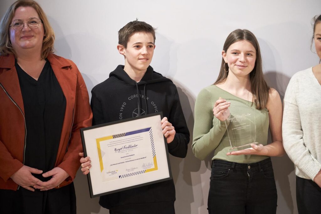 Schüler mit Urkunde und Preis, umgeben von Lehrerin und anderen Personen.