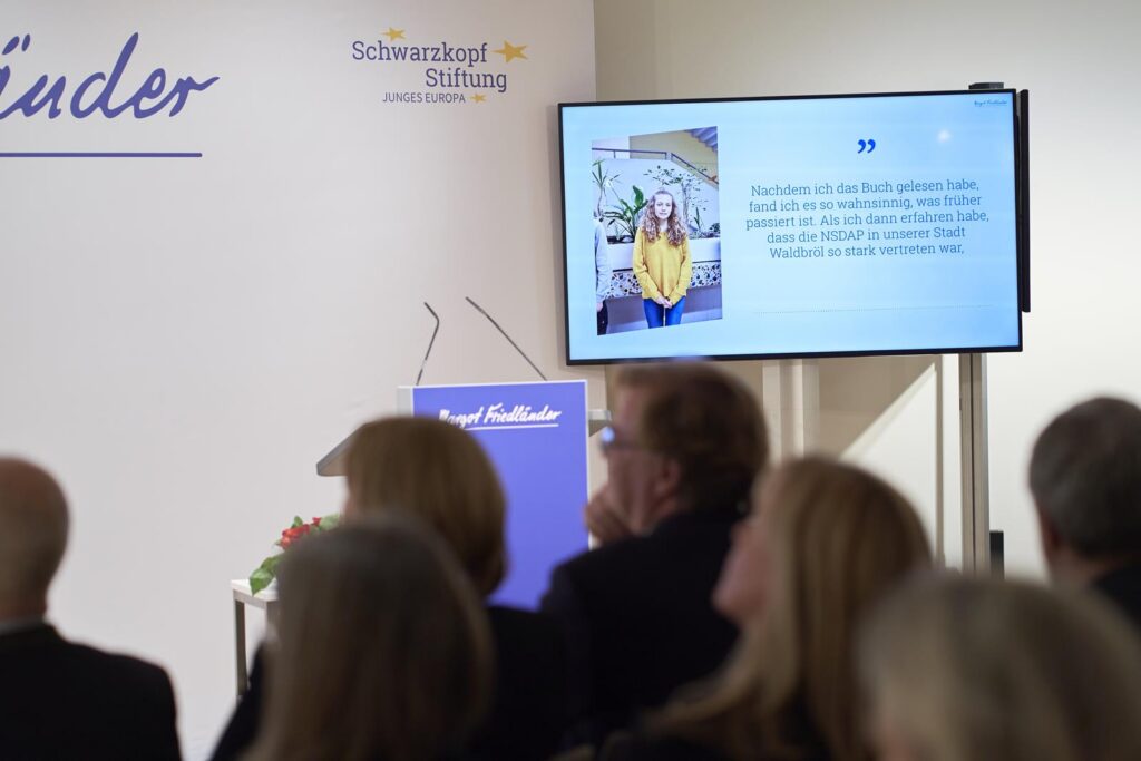 Podium mit Bildschirm und Publikum auf einer Veranstaltungsbühne.