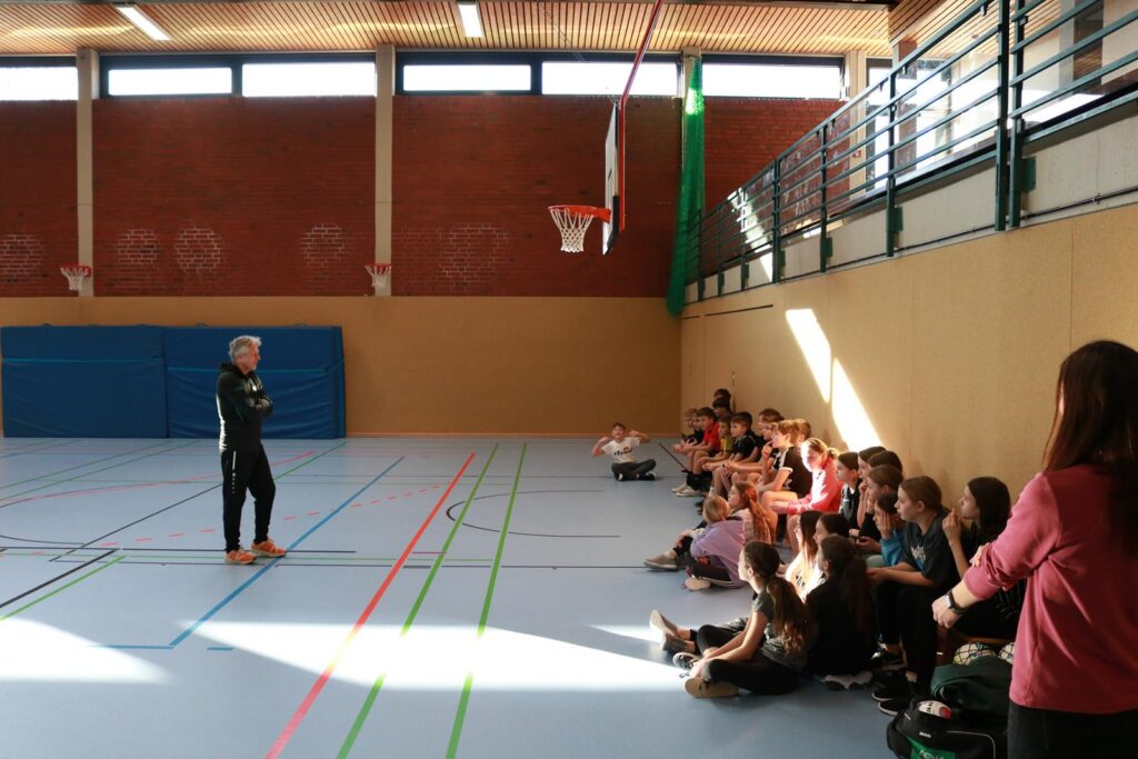 Hallenboden mit Schülern, die an der Wand sitzen, und einem Trainer, der spricht.