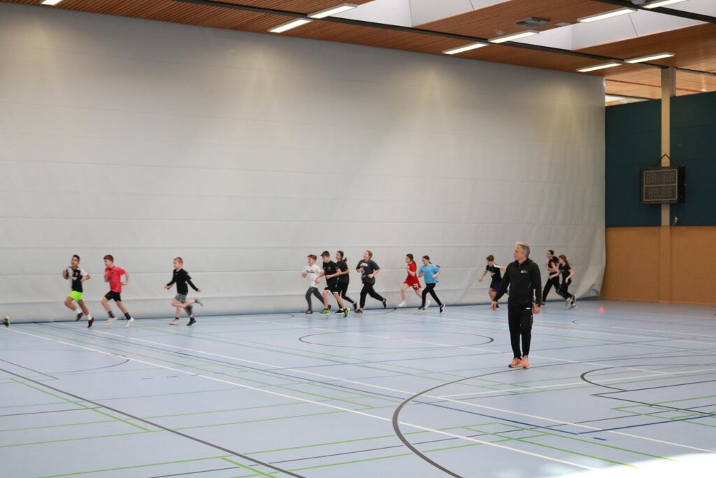Schüler trainieren in einer Sporthalle, einige laufen, andere stehen.
