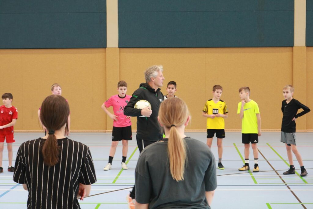 Trainer erklärt Technik im Sportunterricht, Schüler hören aufmerksam zu.