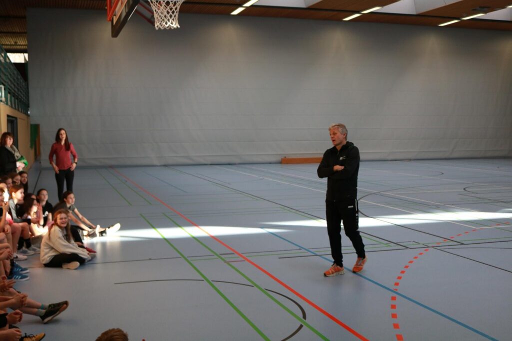 Ein Trainer spricht zu einer Gruppe von Schülern in einer Sporthalle.