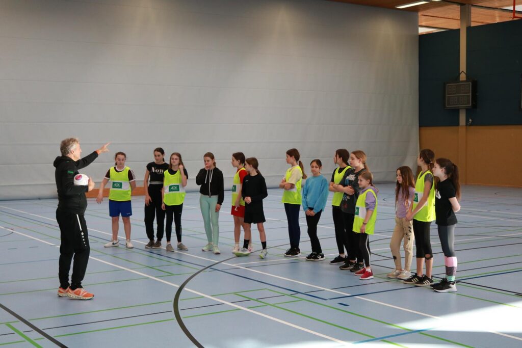 Mädchen in Sportkleidung hören einer Trainerin in einer Halle zu.