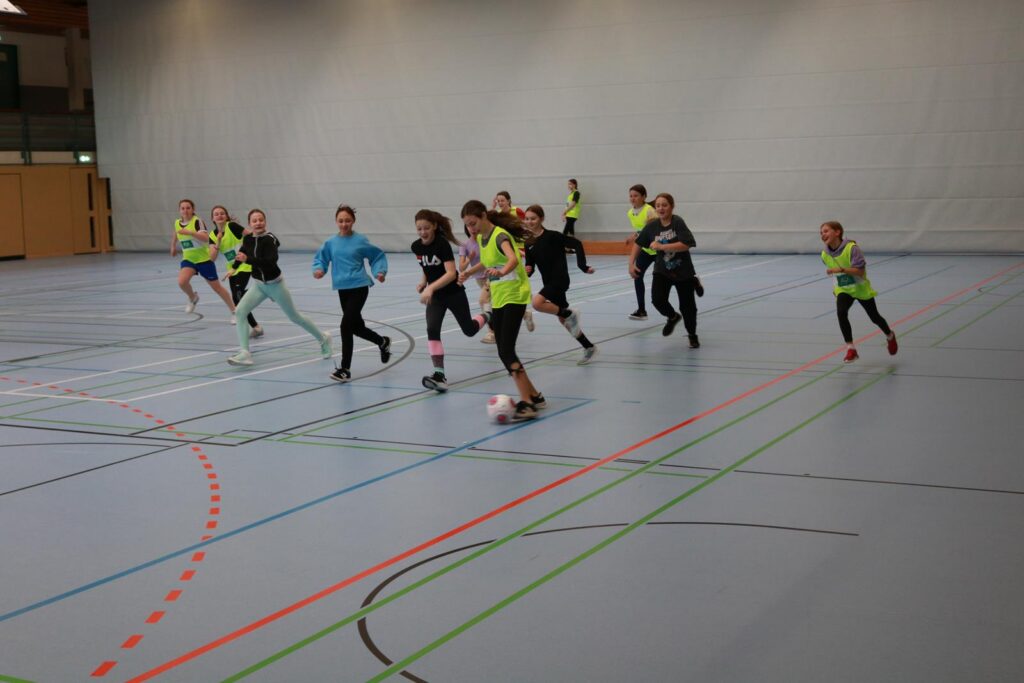 Kinder spielen Fußball in einer Sporthalle. Einige tragen Westen.
