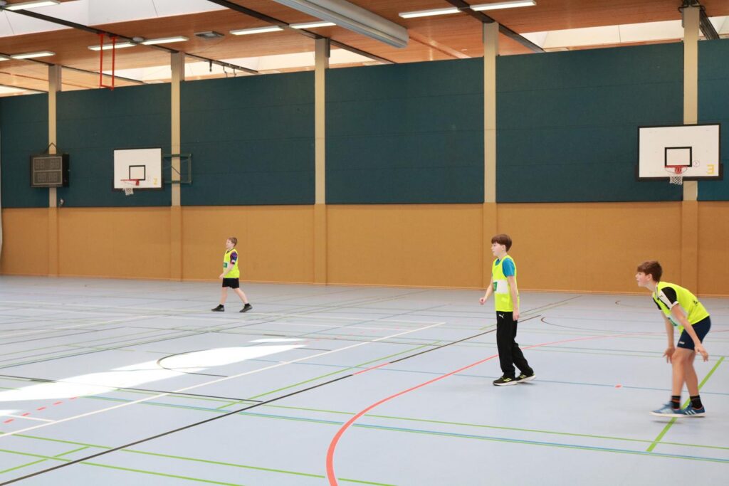 Drei Kinder in Sportkleidung spielen in einer Halle mit Basketballkörben.