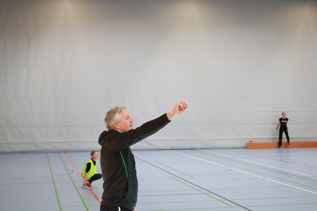 Trainer gibt Anweisungen in einer Sporthalle, Sportler im Hintergrund.