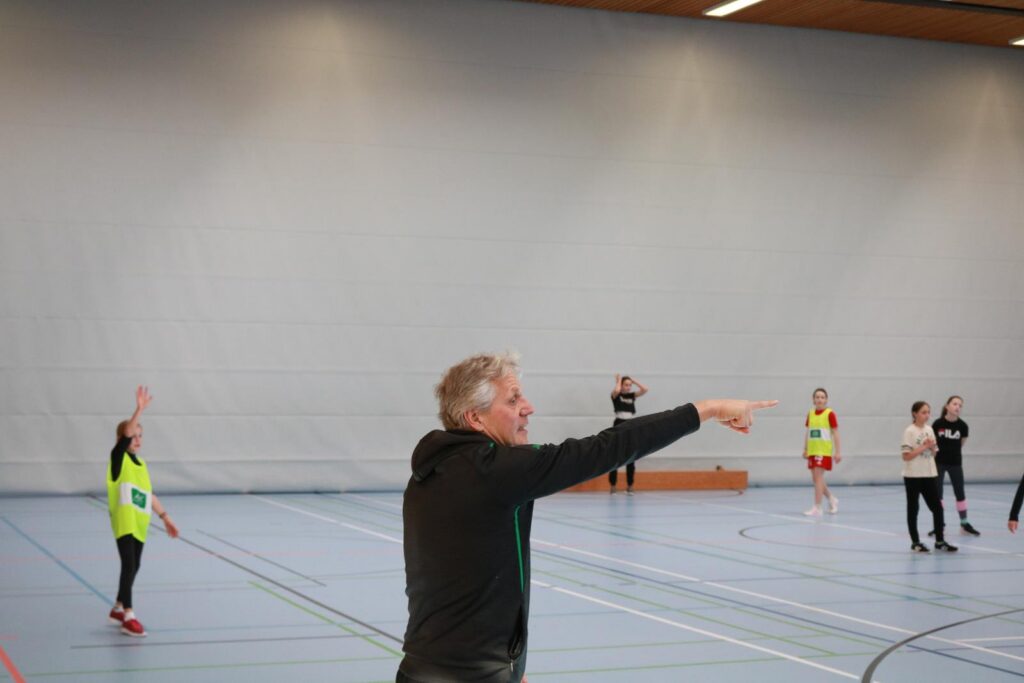 Sporttrainer gibt Anweisungen in einer Sporthalle. Schüler*innen sind aktiv.
