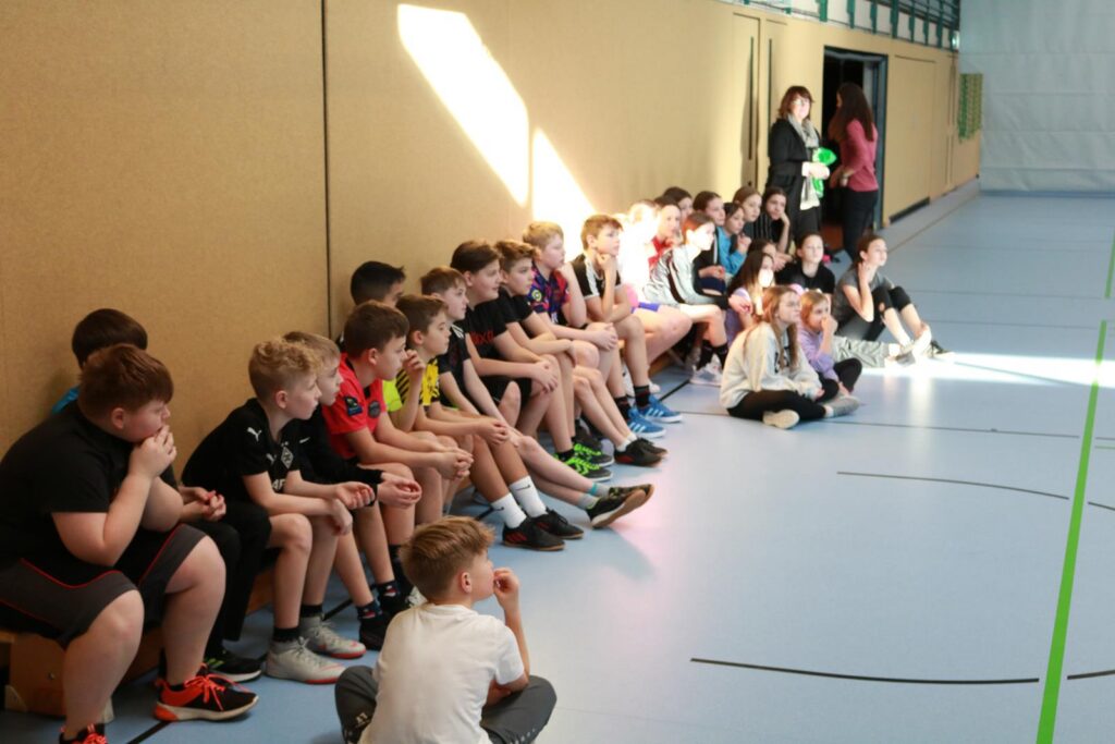 Schüler sitzen in einer Sporthalle, einige schauen nach vorne, andere unterhalten sich.