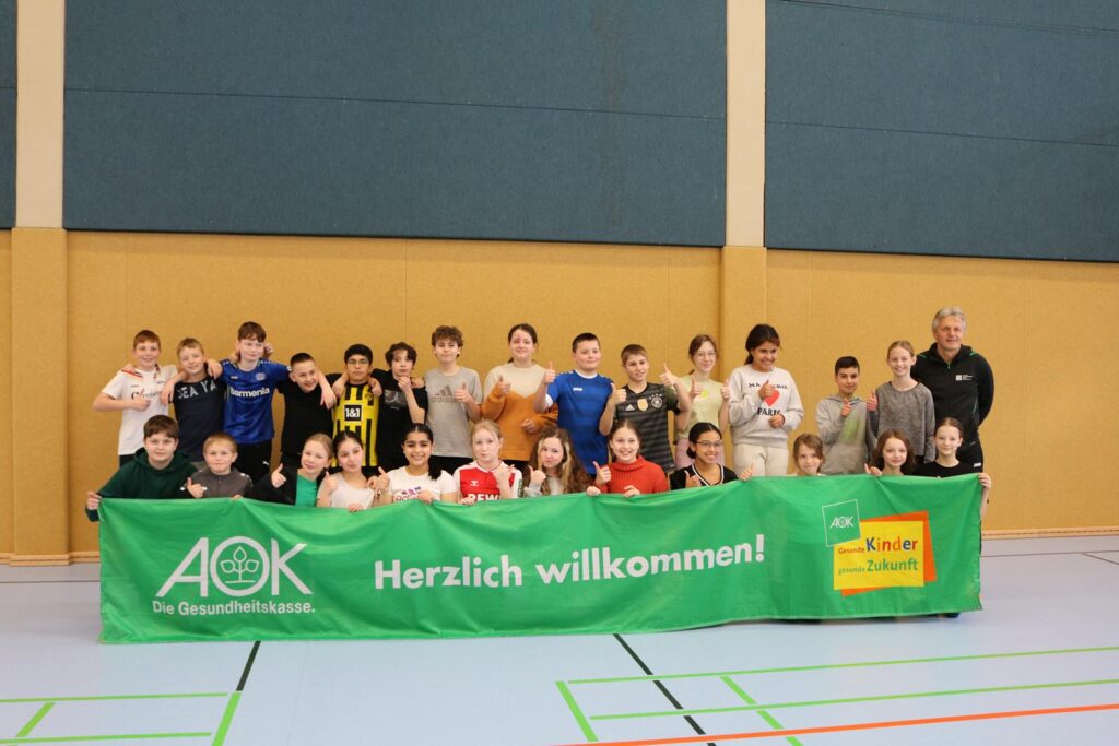 Gruppe von Jugendlichen und Betreuern steht vor einem Banner in einer Sporthalle.