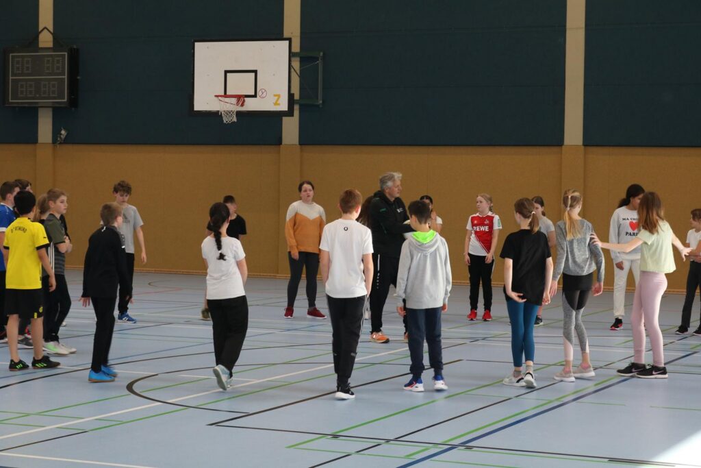 Schülerinnen und Schüler stehen in einer Sporthalle und üben gemeinsam.