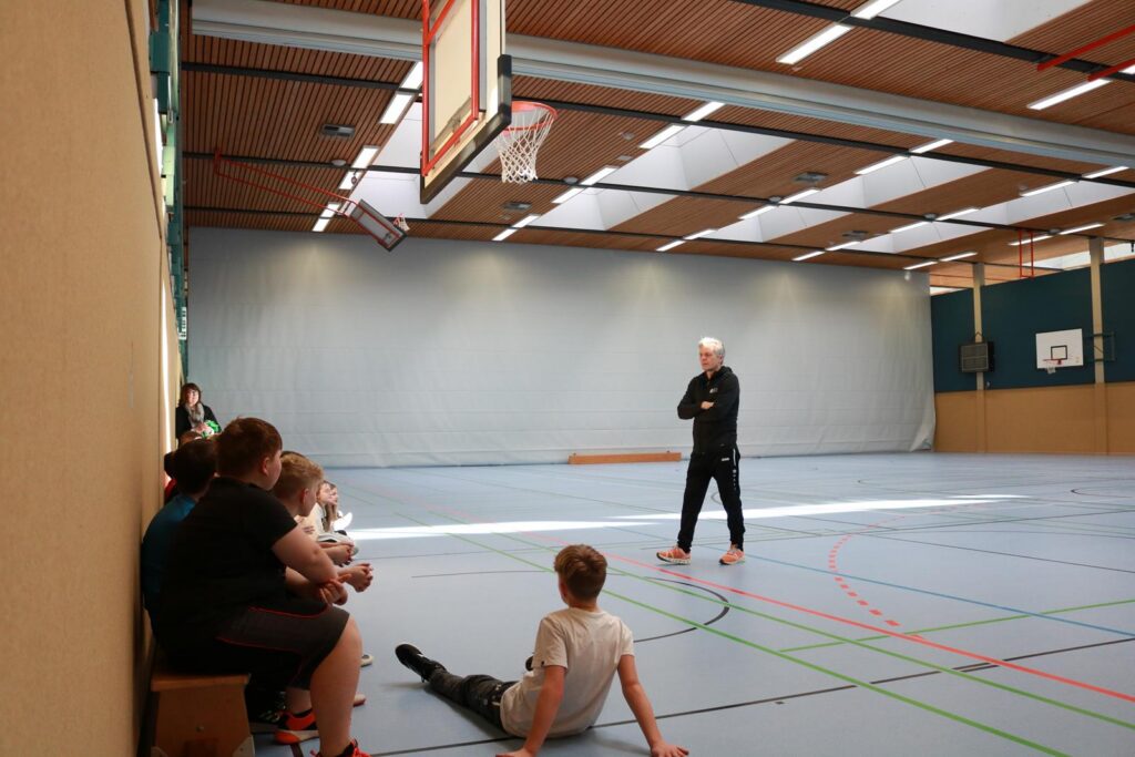 Sporthalle mit Schülern, die an der Wand sitzen, und einem Trainer, der spricht.
