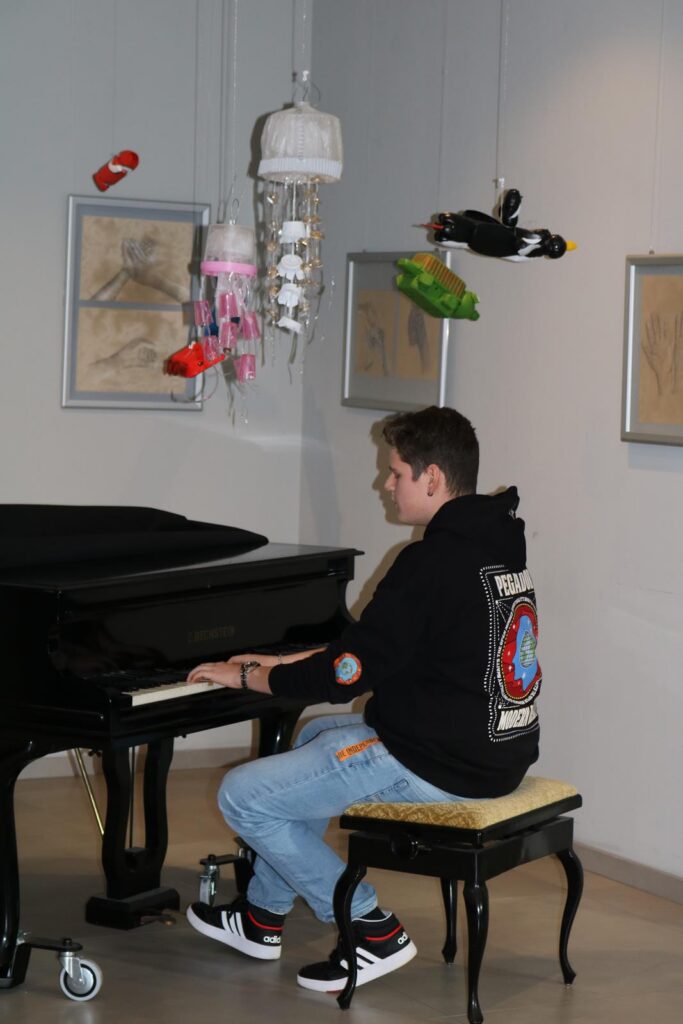 Junge spielt am Klavier, umgeben von hängenden Kunstobjekten und Zeichnungen.