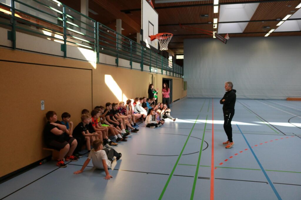 Kinder sitzen an der Wand in einer Sporthalle, Trainer spricht zu ihnen.
