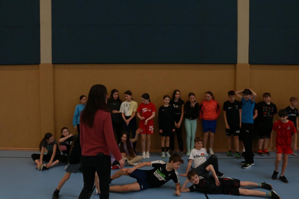 Gruppensituation mit Schülern in einer Sporthalle, einige liegen am Boden.