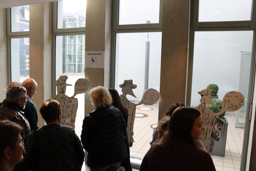 Besucher stehen vor Fenstern mit Figuren und Sprechblasen im Innenraum.