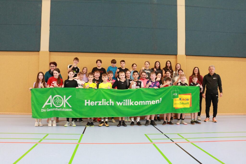 Gruppe von Schülern und Lehrern hält ein Willkommensbanner in einer Sporthalle.