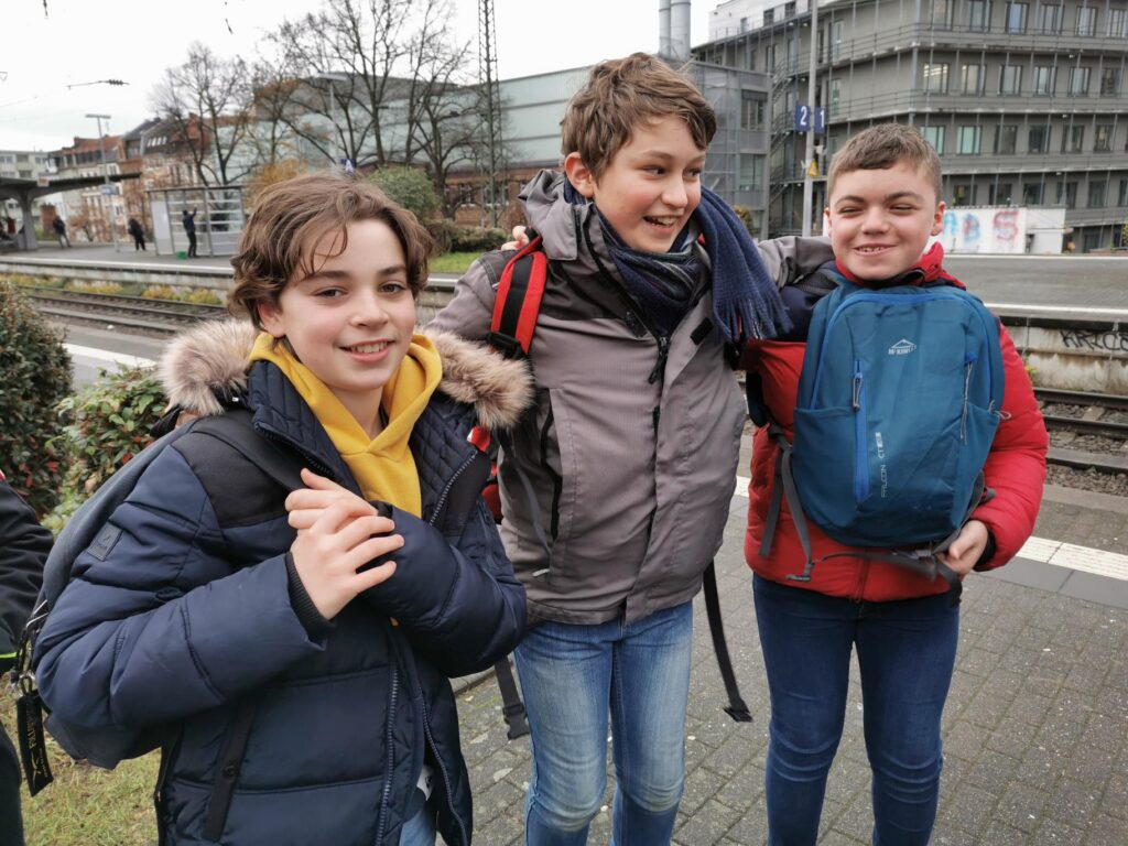 Drei Kinder stehen lächelnd an einer Bahnhaltestelle.