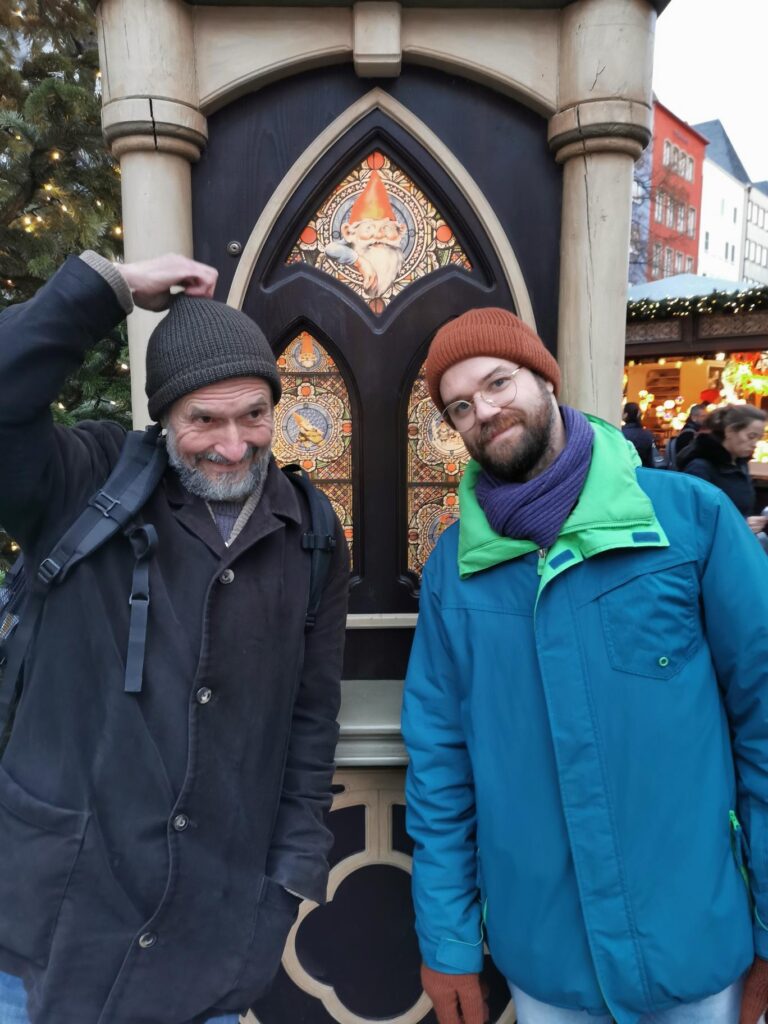 Zwei Männer posieren lächelnd vor einem festlich dekorierten Verkaufsstand.