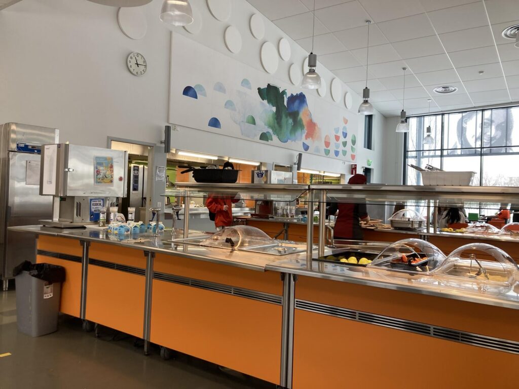 Moderne Schulcafeteria mit bunten Wänden und verschiedenen Essensstationen.