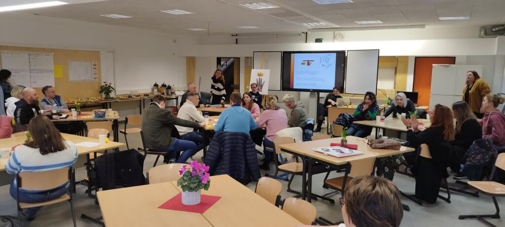 Gruppendiskussion in einem Klassenzimmer mit Tischen und Präsentationsbildschirm.