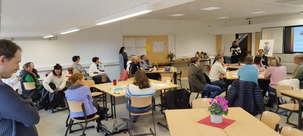 Gruppensitzung mit Teilnehmern in einem Raum, Tische und Präsentation im Hintergrund.