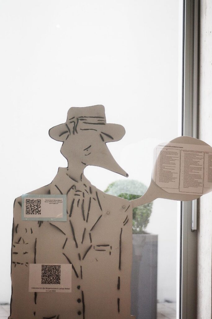 Papierfigur eines Mannes mit Hut und langem Schnabel vor einer Glaswand.