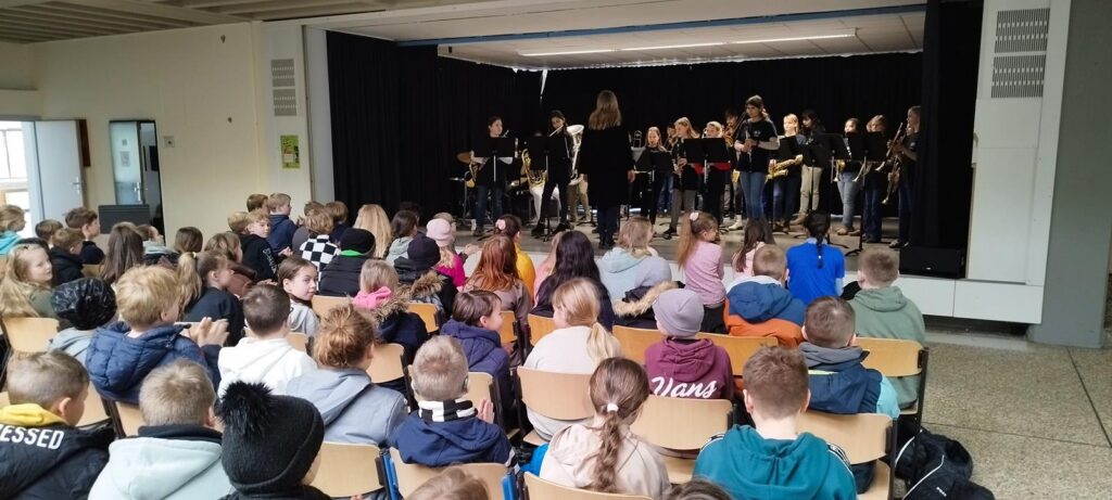 Schüler sitzen vor einer Band auf der Bühne in einem Veranstaltungsraum.