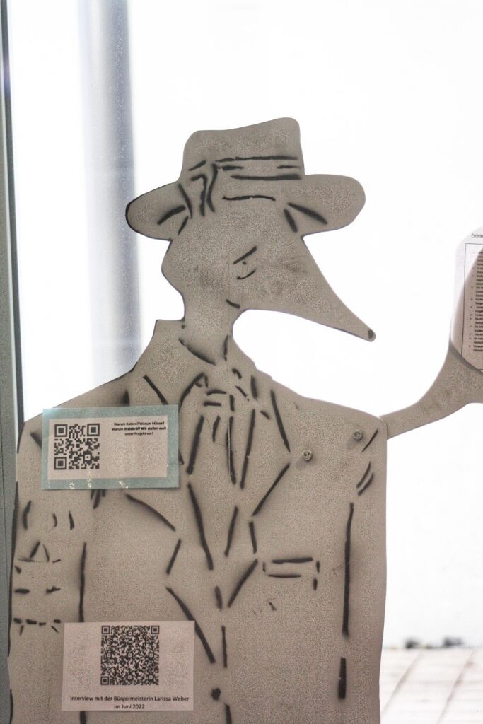 Skelettartige Figur eines Mannes mit Hut und Anzug steht an einem Fenster.