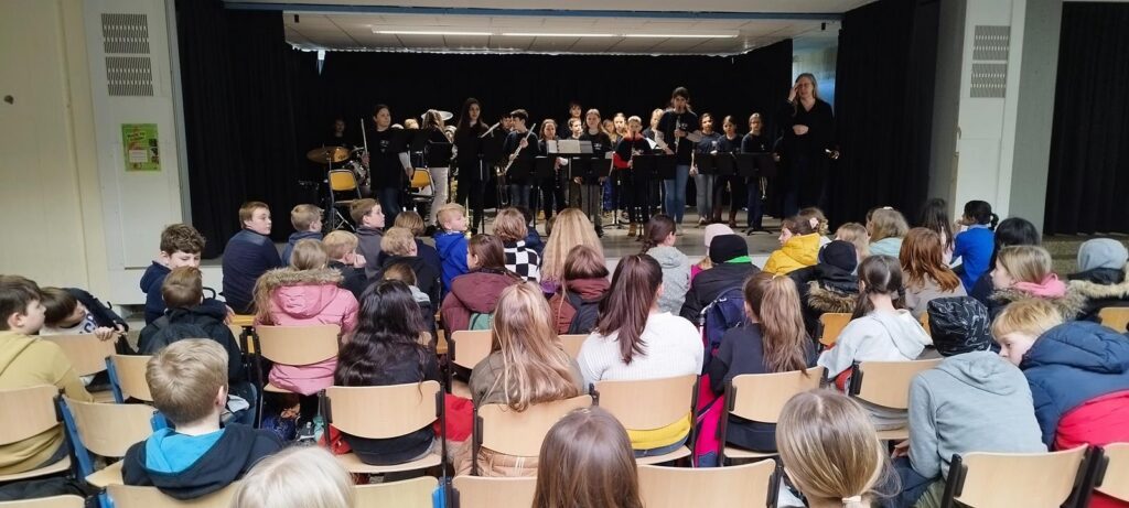 Schüler sitzen auf Stühlen und schauen auf ein Orchester auf der Bühne.
