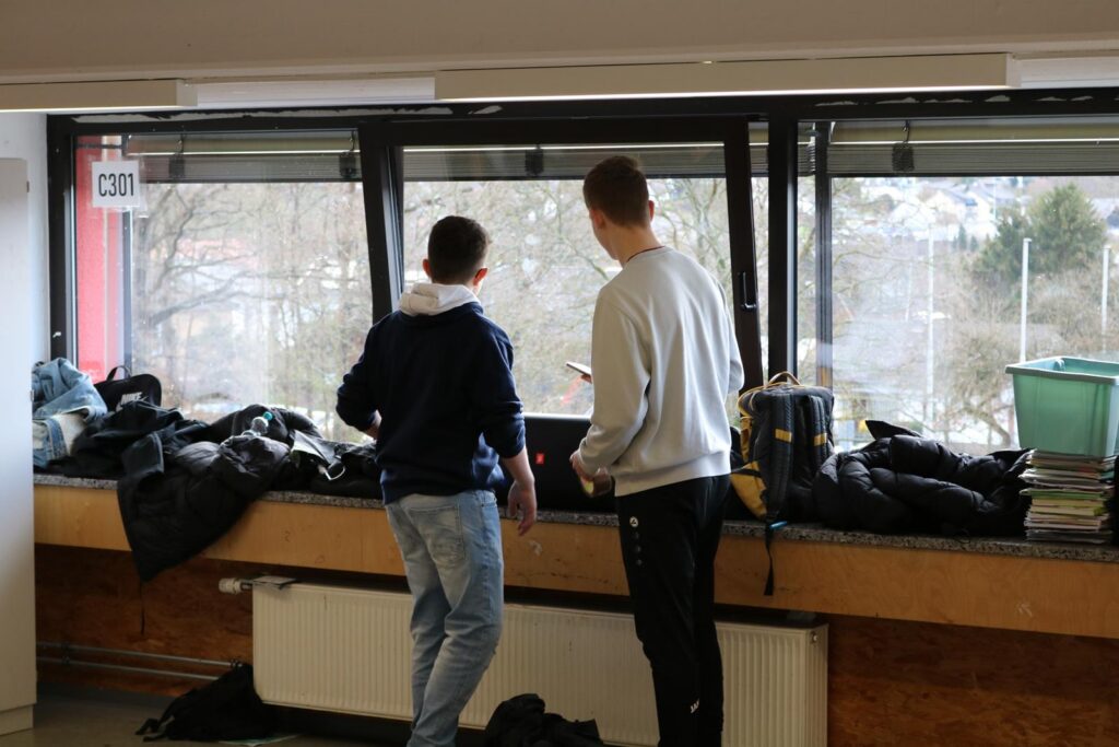 Zwei Schüler stehen am Fenster und schauen nach draußen. Jacken liegen auf der Fensterbank.