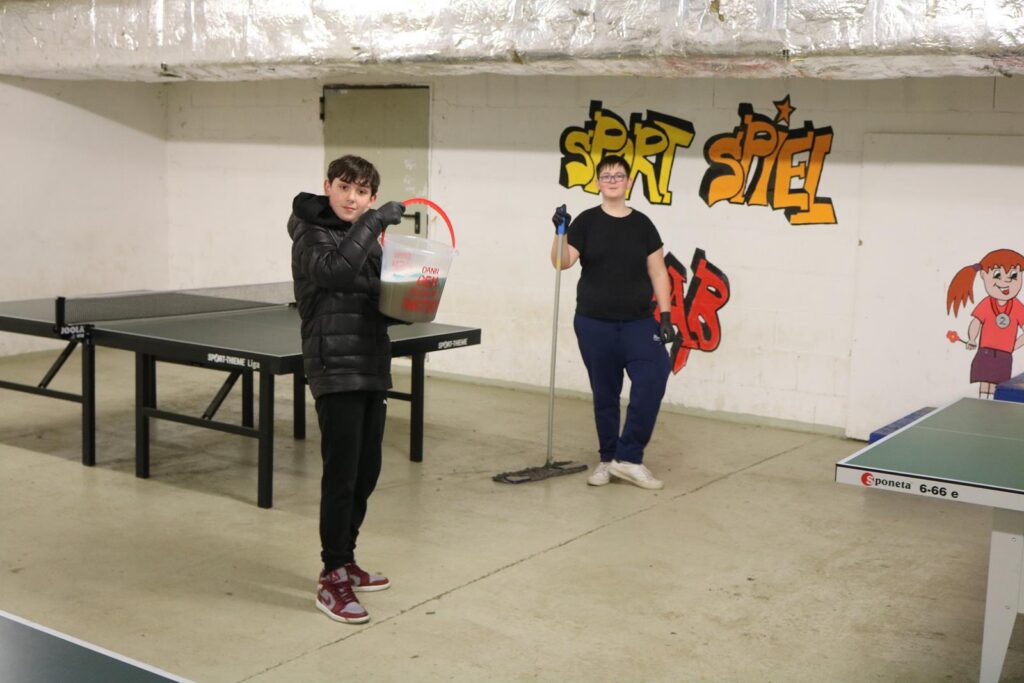Zwei Jungen in einem Raum mit Tischtennisplatten, einer hält einen Eimer, der andere moppt.