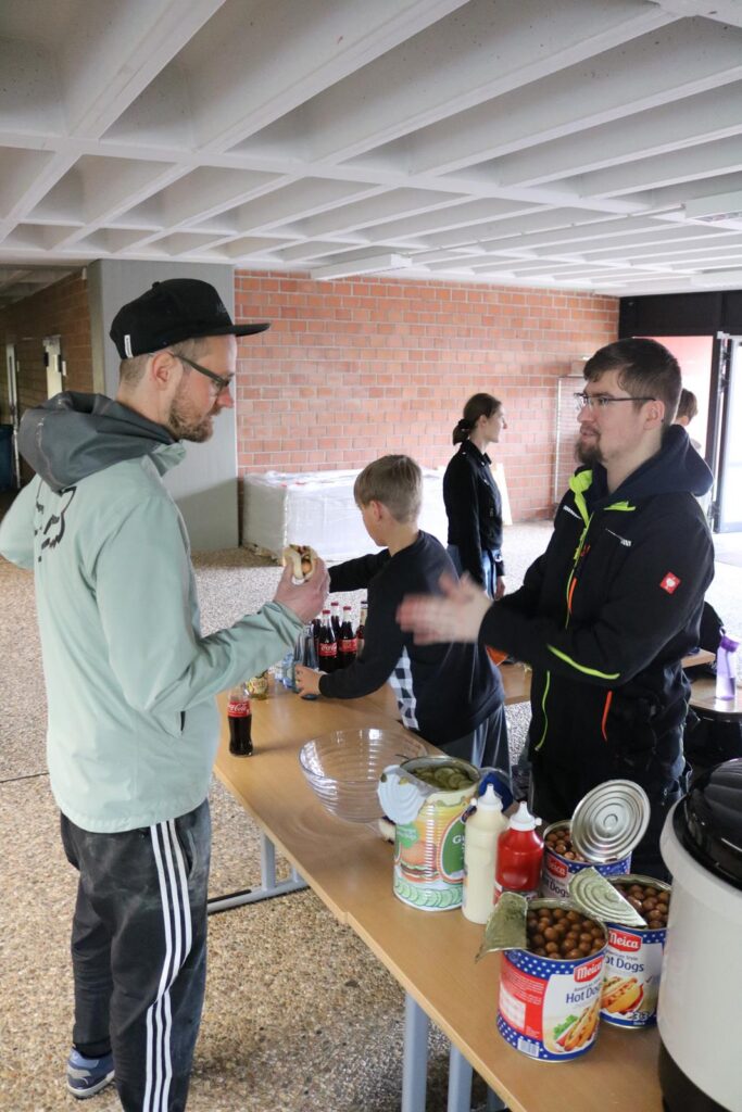 Tisch mit Essensständen, zwei Personen im Gespräch, Kinder im Hintergrund.