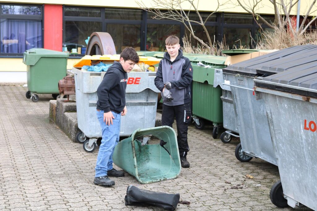 Zwei Jungen stehen neben Abfallcontainern und untersuchen einen grünen Behälter.