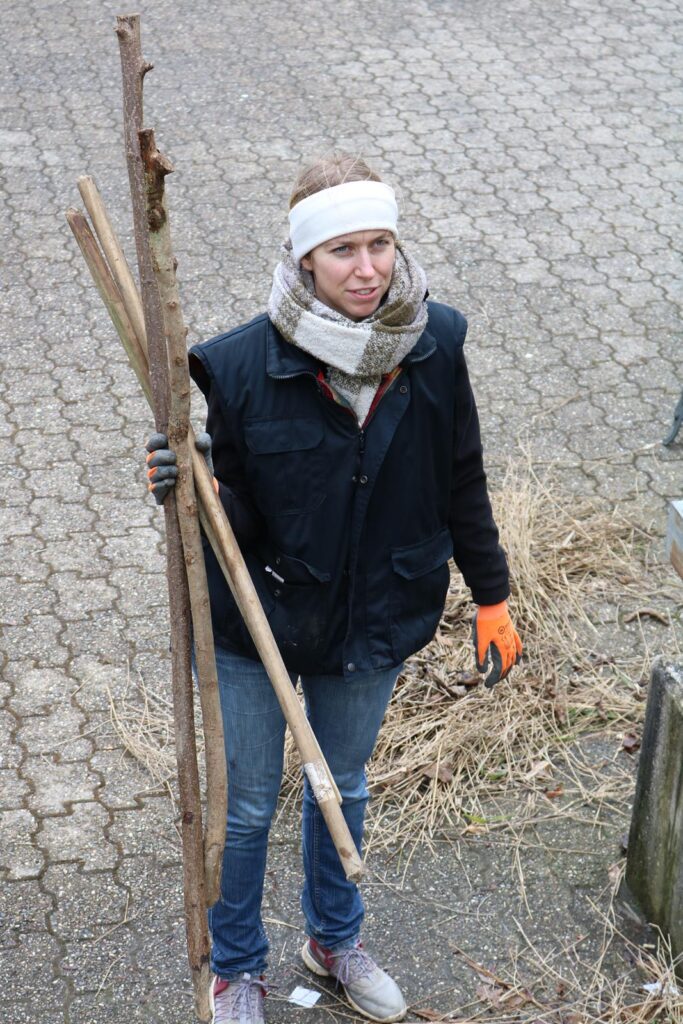Frau hält zwei Holzstöcke und trägt eine Jacke sowie einen Schal.