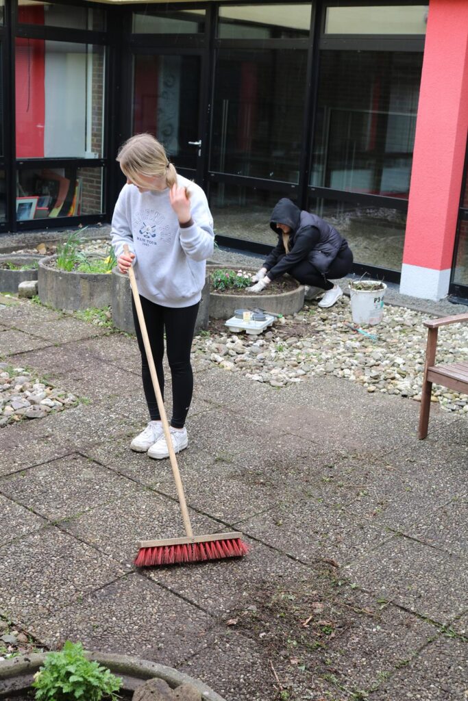 Schülerinnen fegen und bepflanzen einen schulischen Garten.