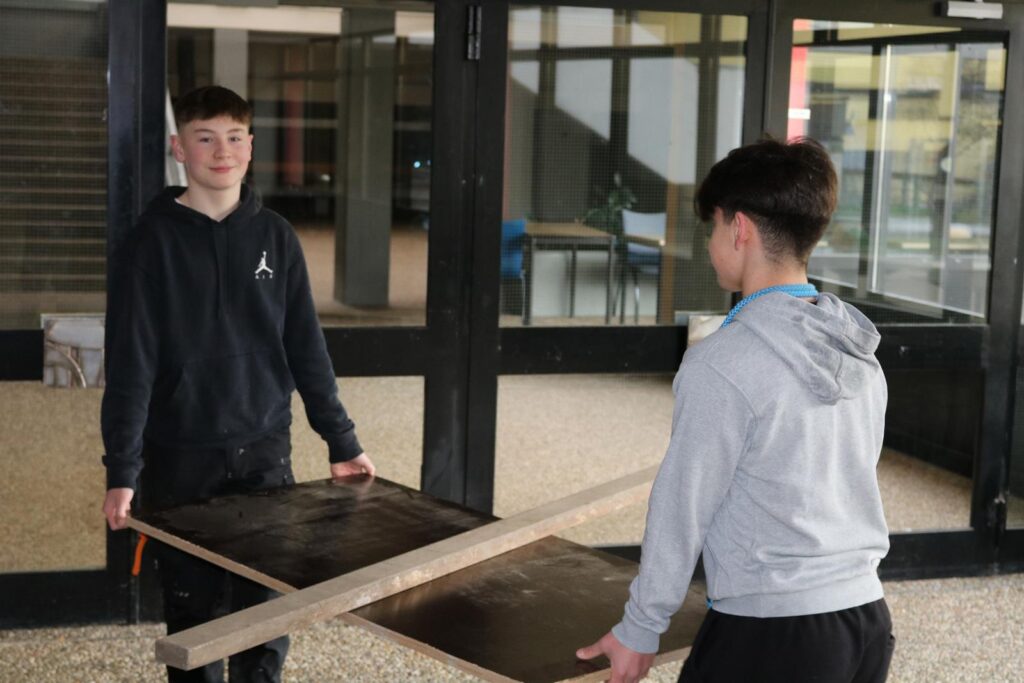 Zwei Schüler tragen gemeinsam eine Tischplatte in einer Schule.