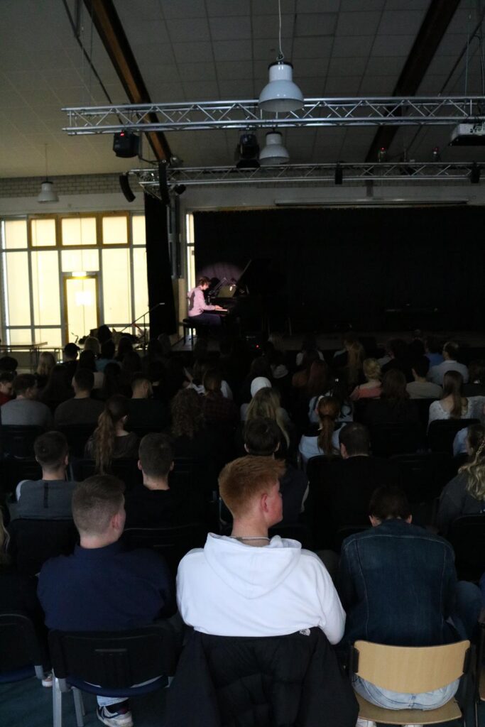 Publikum in einem Saal bei einer Veranstaltung. Bühne im Hintergrund sichtbar.