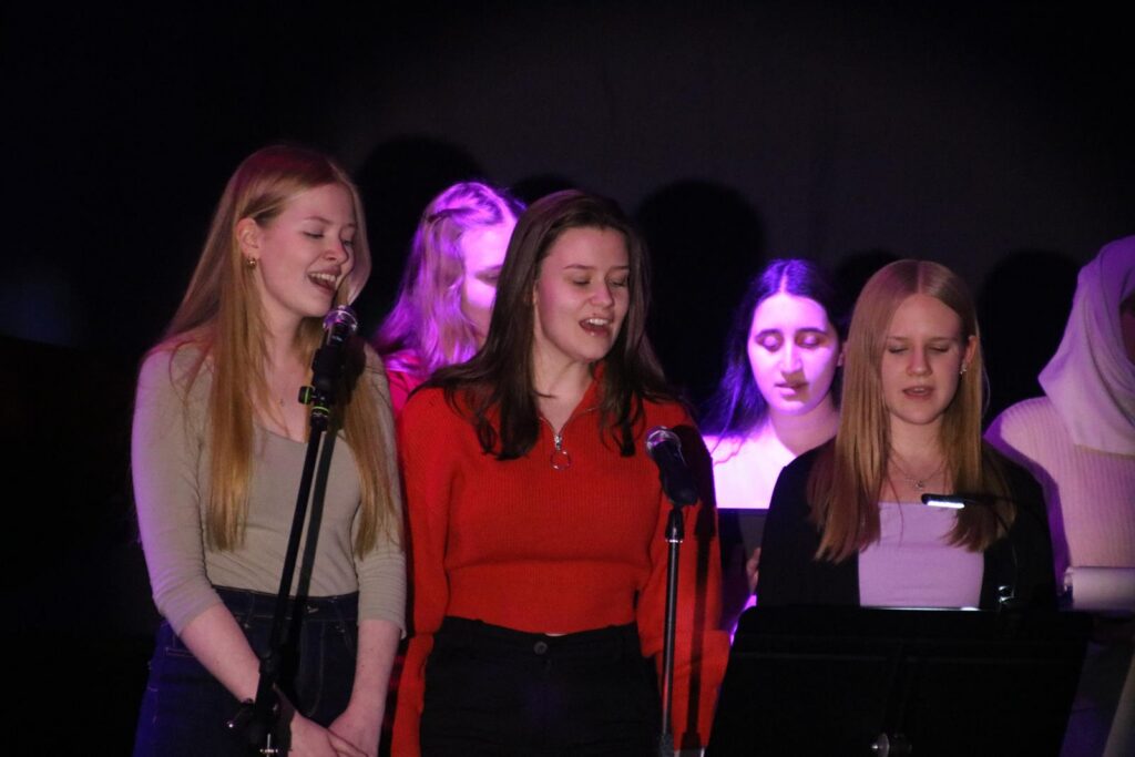 Drei Sängerinnen stehen am Mikrofon und singen vor einer Gruppe.