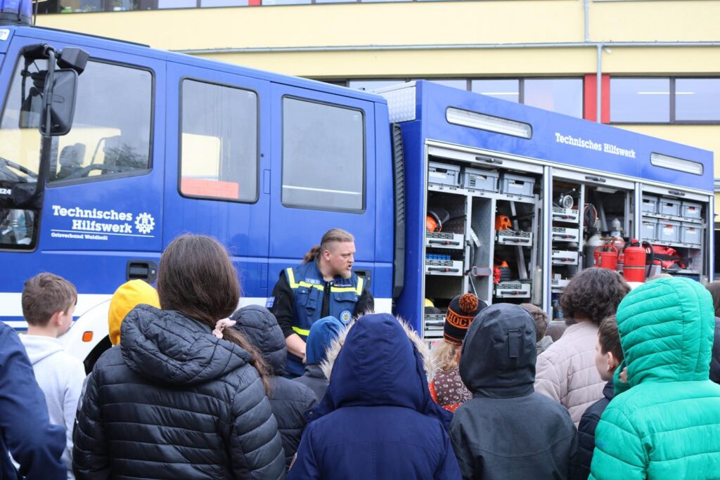 Feuerwehrmann erklärt Ausrüstung vor einem Löschfahrzeug, Schüler hören zu.