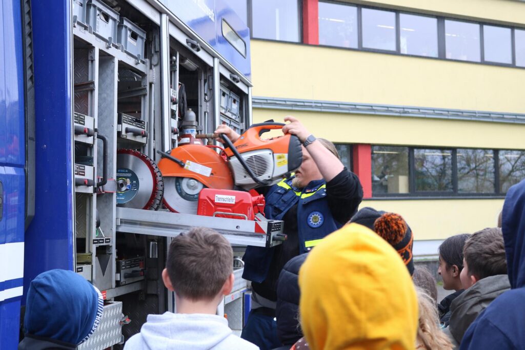 Feuerwehrmann zeigt Ausrüstung einer Feuerwehraufschnittmaschine. Schüler beobachten interessiert.