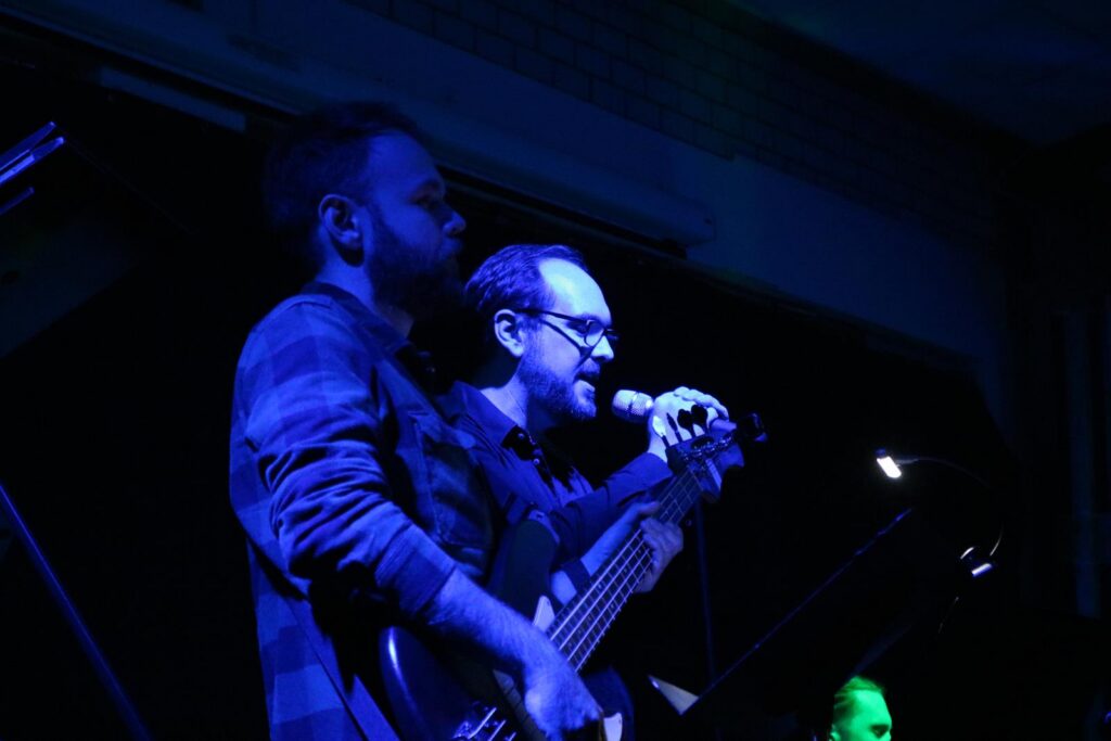 Zwei Musiker stehen auf der Bühne und spielen Instrumente in blauer Beleuchtung.