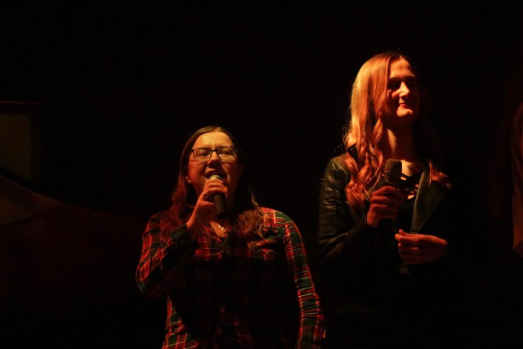 Zwei Frauen stehen auf einer Bühne und singen in einem schwach beleuchteten Raum.