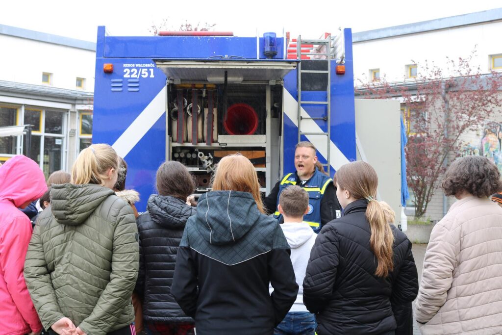Besuchergruppe hört einem Feuerwehrmann vor einem Feuerwehrauto zu.