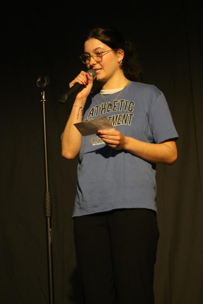 Eine Person steht am Mikrofon und spricht, trägt ein blaues T-Shirt.