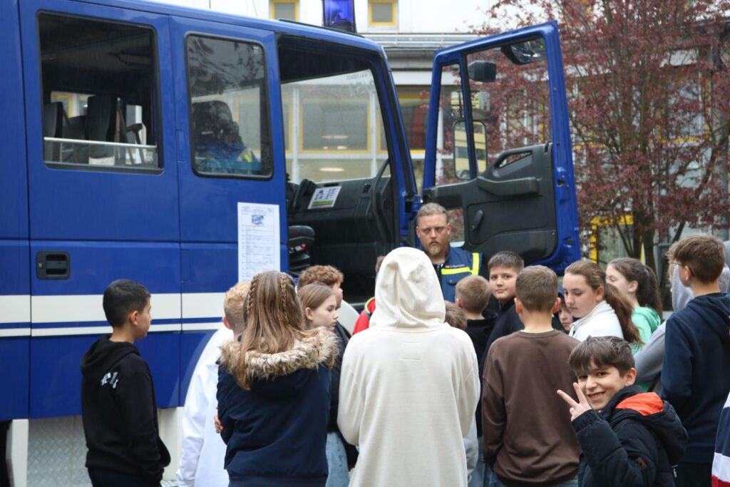 Schüler stehen um ein Polizeifahrzeug und hören einem Beamten zu.